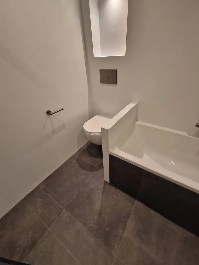 Hangtoilet naast het ligbad met verlichte nis en grijze vloertegels — badkamerrenovatie in Roden door BontBouw