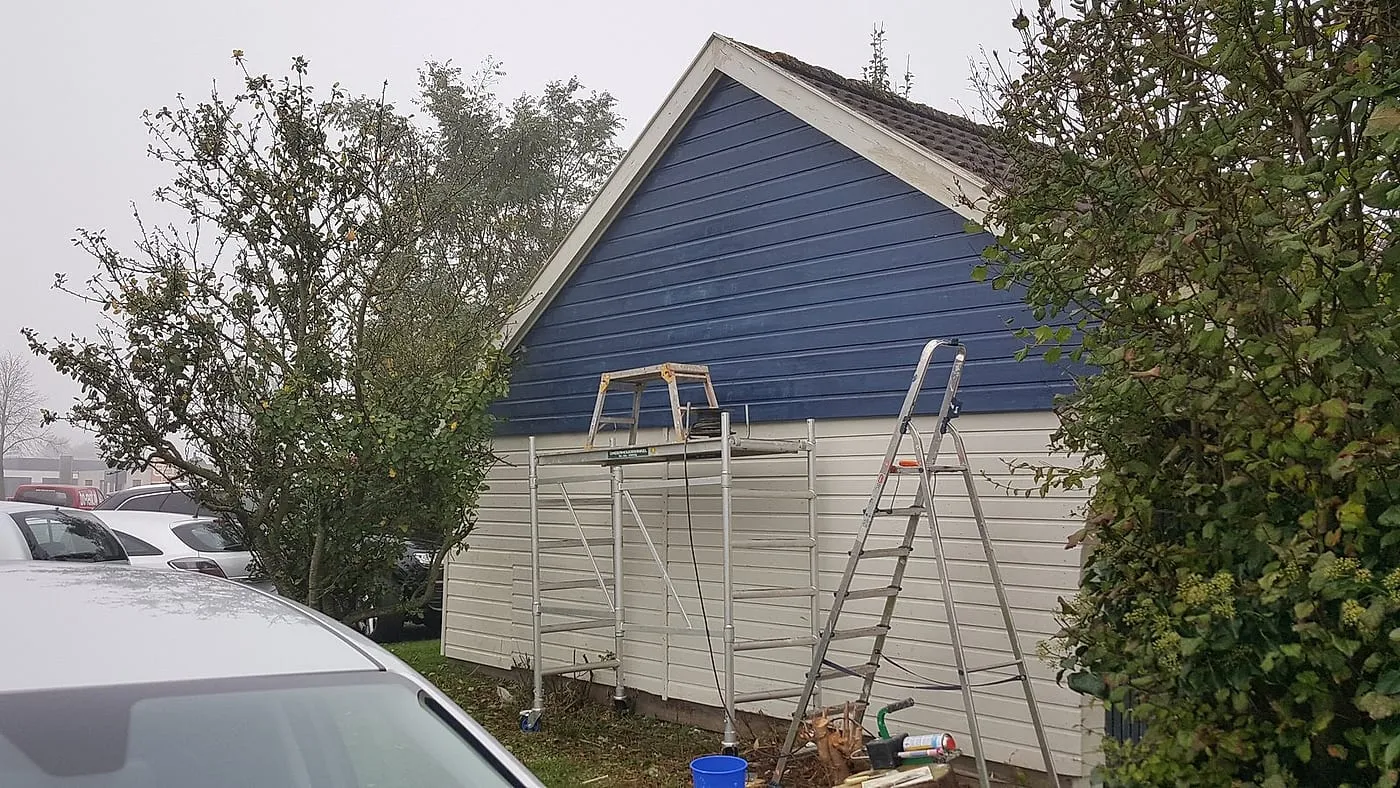 Verweerde blauwe houten topgevel van de garage met rolsteiger vóór het werk — schilderwerk in Euvelgunne door BontBouw